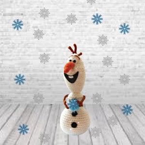 以下が含まれることがあります： 映画アナと雪の女王のキャラクター、オラフの編みぐるみおもちゃ。青い雪玉を持っています。おもちゃは白い木製の表面に立っており、白いレンガの壁が背景になっています。おもちゃの周りに、グレーとブルーの雪の結晶がいくつか散らばっています。
