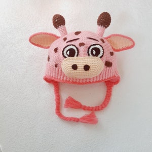 Könnte beinhalten: Ein gehäkelter Giraffenhut in Rosa und Braun mit großen Augen, einer beigen Schnauze und braunen Flecken. Der Hut hat Ohrenklappen und geflochtene Bänder mit Quasten. Der Hut ist zum Tragen auf dem Kopf bestimmt.