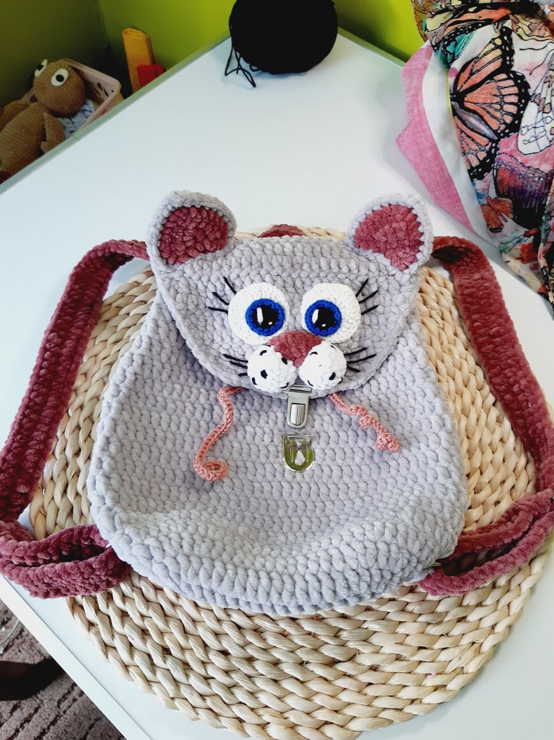 May include: A gray crocheted cat backpack with pink ears and blue eyes. The backpack has a metal clasp closure and is sitting on a woven straw mat.