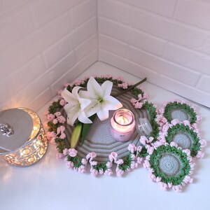 K&ouml;nnte beinhalten: Ein Set geh&auml;kelter Untersetzer mit einem gr&uuml;n-grauen Muster und rosa Einfassung. Eine wei&szlig;e Lilie und eine Kerze liegen auf einem gr&ouml;&szlig;eren geh&auml;kelten Untersetzer.