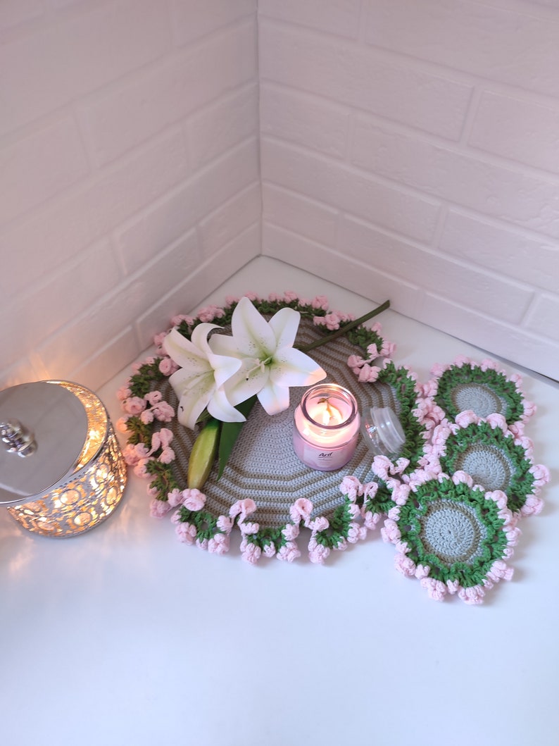 K&ouml;nnte beinhalten: Ein Set geh&auml;kelter Untersetzer mit einem gr&uuml;n-grauen Muster und rosa Einfassung. Eine wei&szlig;e Lilie und eine Kerze liegen auf einem gr&ouml;&szlig;eren geh&auml;kelten Untersetzer.