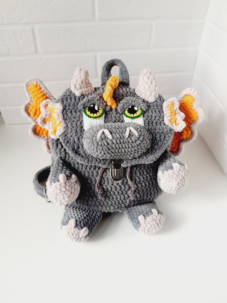 May include: Gray crocheted dragon backpack with orange wings, pink feet, and green eyes. The backpack has a zipper closure and a small handle on top.