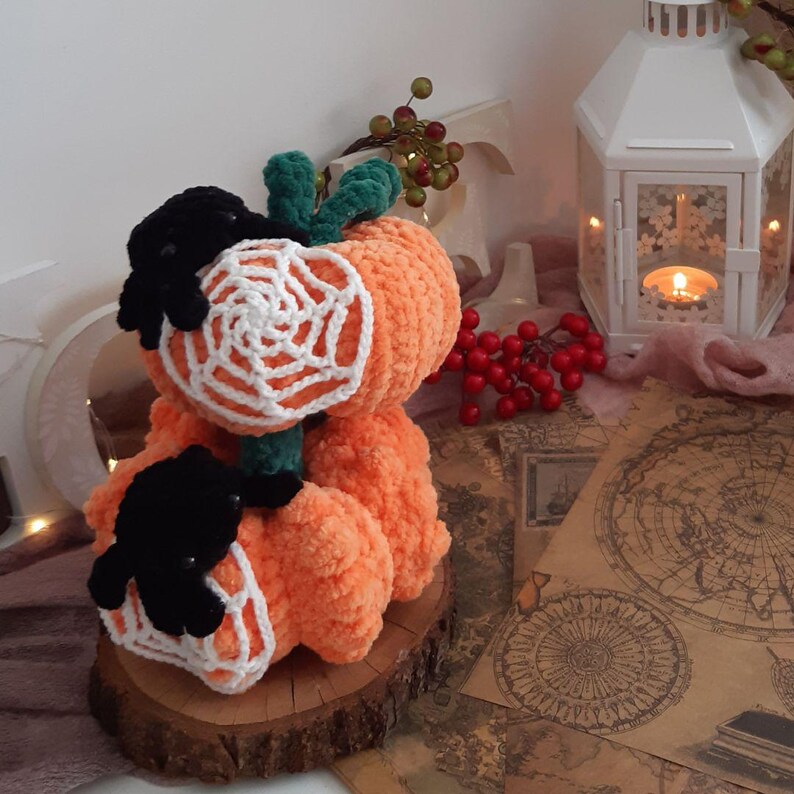 May include: Three crocheted orange pumpkins with black spiders and white crocheted spiderwebs. The pumpkins are stacked on a wooden slice.