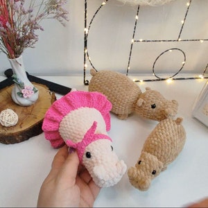 Puede incluir: Tres juguetes de hipopótamo de crochet. Uno es rosa con un tutú rosa, uno es marrón y uno es marrón con la nariz rosa.