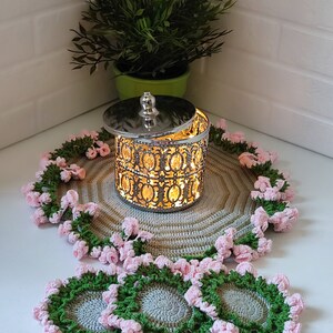 K&ouml;nnte beinhalten: Set aus vier geh&auml;kelten Untersetzern mit einem rosa-gr&uuml;nen Blumenmuster. Die Untersetzer sind aus grauem Garn gefertigt und haben ein erhabenes Blumenmuster. Die Untersetzer werden auf einem geh&auml;kelten Platzdeckchen mit einem silbernen Metallbeh&auml;lter in der Mitte pr&auml;sentiert.