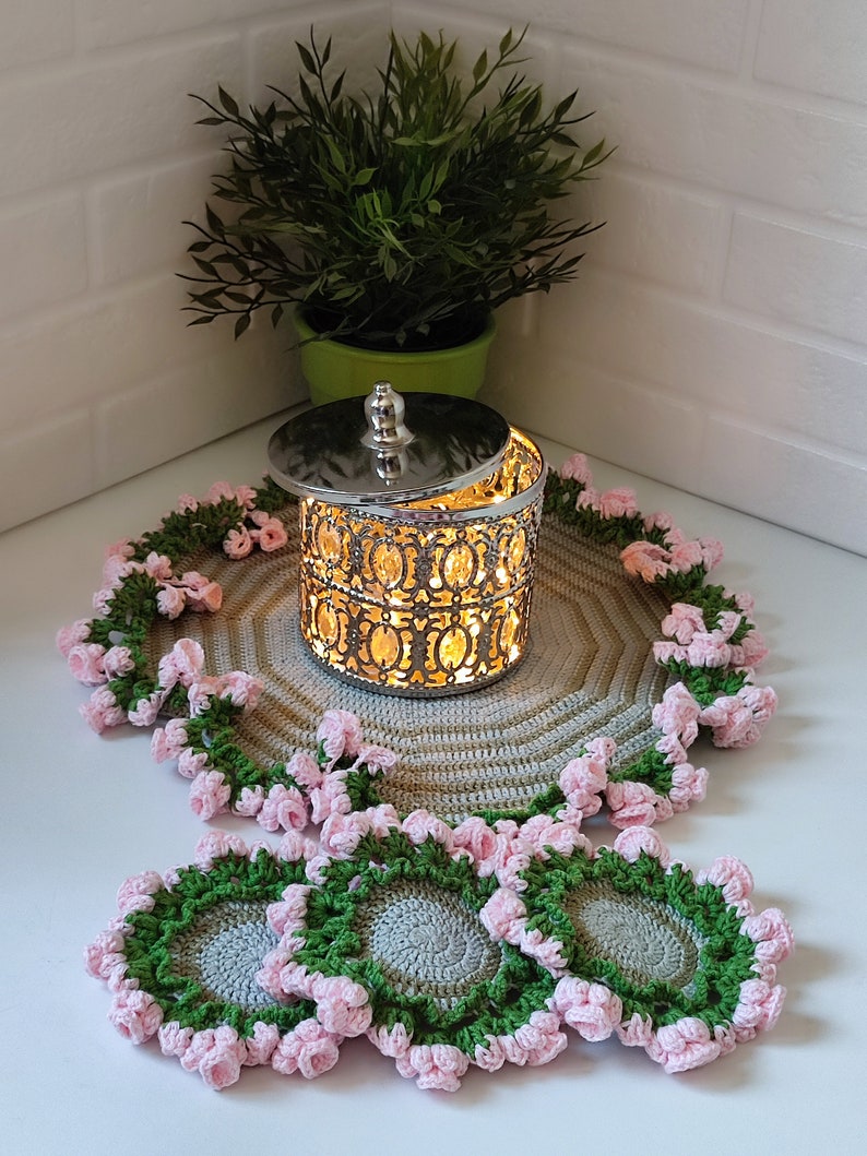 K&ouml;nnte beinhalten: Set aus vier geh&auml;kelten Untersetzern mit einem rosa-gr&uuml;nen Blumenmuster. Die Untersetzer sind aus grauem Garn gefertigt und haben ein erhabenes Blumenmuster. Die Untersetzer werden auf einem geh&auml;kelten Platzdeckchen mit einem silbernen Metallbeh&auml;lter in der Mitte pr&auml;sentiert.