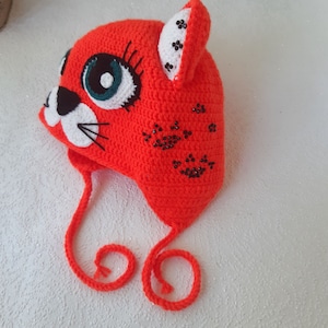 Op de afbeelding: Oranje gehaakte muts met een kattengezicht en zwarte kralen. De muts heeft een lange strik die onder de kin kan worden gebonden.