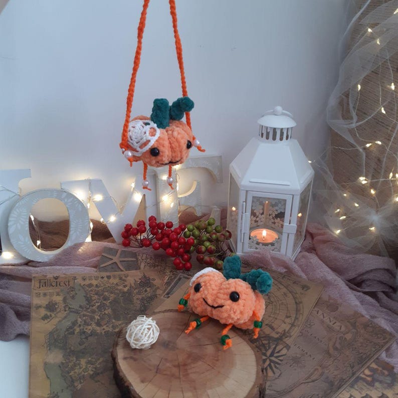 May include: Two crocheted pumpkin plushies, one orange with green leaves and a white crocheted flower, the other orange with green leaves. The pumpkin plushies are sitting on a wooden surface with a map and a white lantern.