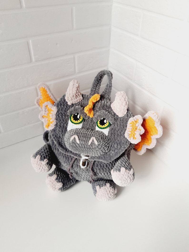 May include: Gray crocheted dragon backpack with orange wings and yellow horns. The backpack has a zipper closure and green eyes.