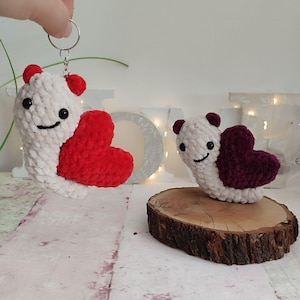 May include: Two handmade crochet heart-shaped creatures. One is a keychain, the other is on a wooden slice. The creatures are white with red hearts, small red ears, and smiling faces. The second creature has a burgundy heart.