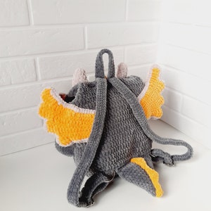 May include: Gray crocheted backpack shaped like a dragon with orange and pink wings. The backpack has adjustable straps.