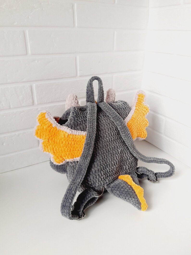 May include: Gray crocheted backpack shaped like a dragon with orange and pink wings. The backpack has adjustable straps.