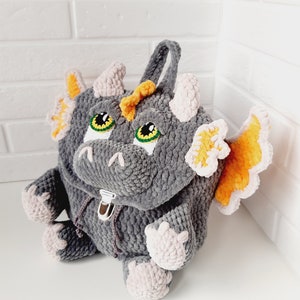 May include: A gray crocheted dragon backpack with orange and pink accents. The backpack has a zipper closure and a handle on top.