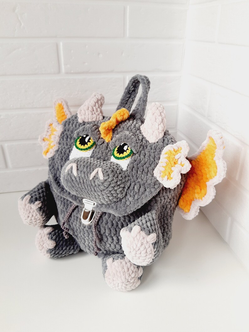 May include: A gray crocheted dragon backpack with orange and pink accents. The backpack has a zipper closure and a handle on top.