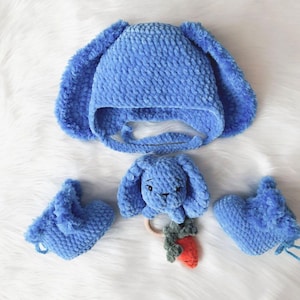 May include: A blue crochet baby set featuring a bunny hat with long ears, a bunny rattle with a carrot, and matching booties. The set is made of soft, plush material, perfect for a newborn or baby shower gift.