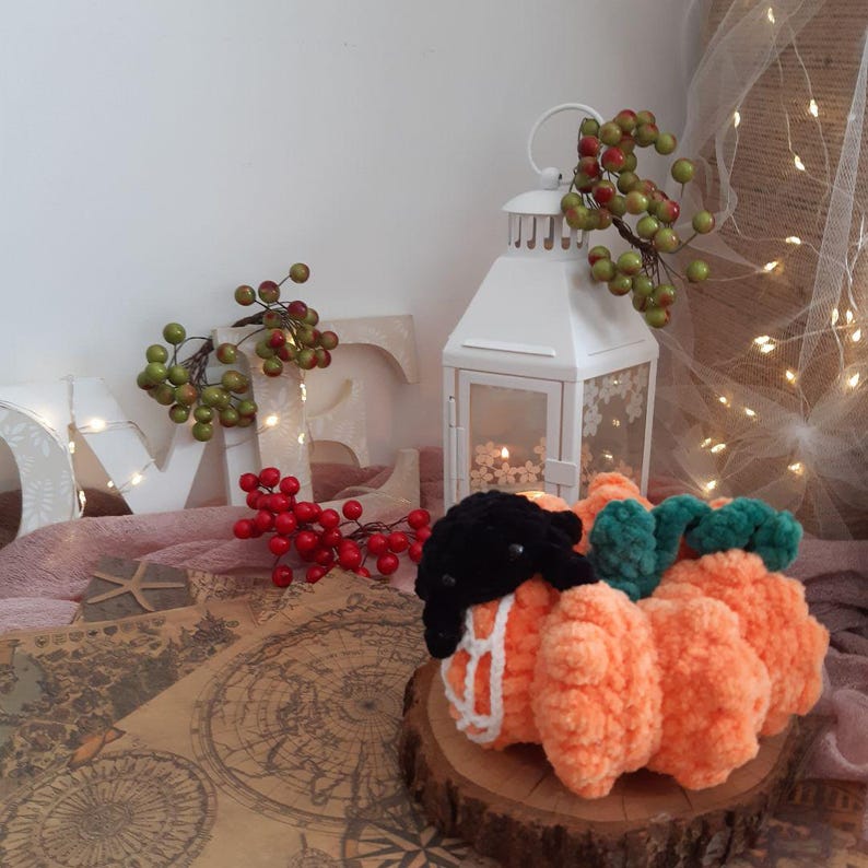 May include: A crocheted orange pumpkin with a black spider on top. The pumpkin has a green stem and white web details. The pumpkin is sitting on a wooden slice.