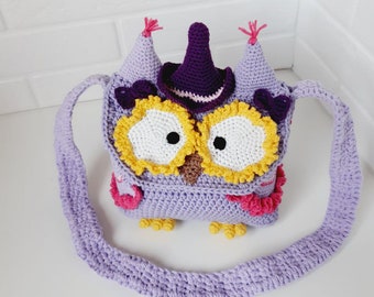 May include: A purple crocheted owl-shaped bag with a purple witch hat, yellow eyes, and a pink and purple ruffle. The bag has a long purple strap.