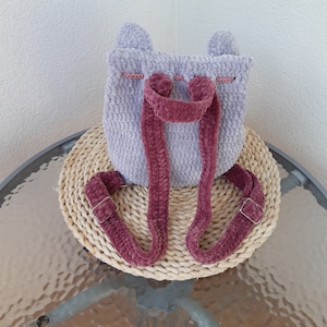 May include: A small purple crocheted backpack with pink straps and bunny ears. The backpack is sitting on a woven mat.