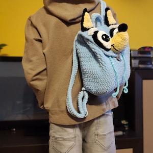 May include: A light blue crocheted backpack with a cartoon dog face. The backpack has a drawstring closure and a small metal buckle. The dog face has yellow ears, black eyes, and a white nose.