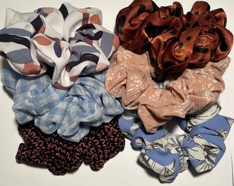 May include: A collection of six colorful hair scrunchies in various patterns and fabrics. The scrunchies include a blue and white checkered pattern, a floral print, a brown and white patterned scrunchie, a solid brown scrunchie, a blue and white floral scrunchie, and a black and red patterned scrunchie.