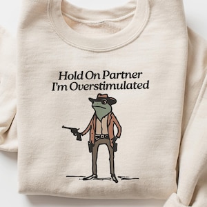 May include: Cream-colored sweatshirt with the text "Hold On Partner I'm Overstimulated." The graphic features a frog dressed as a cowboy holding a gun. The sweatshirt has long sleeves.