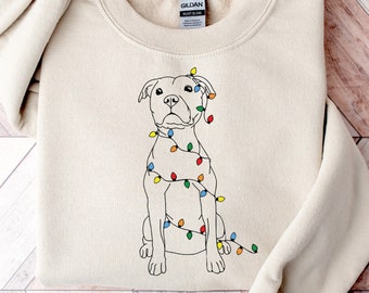 May include: A beige sweatshirt with a white outline drawing of a dog wearing Christmas lights. The dog is sitting and looking to the left. The sweatshirt has the word "Gildan" printed on the tag.