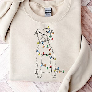 May include: A beige sweatshirt featuring a white outline drawing of a dog wearing Christmas lights. The dog is sitting and looking to the left. The sweatshirt has the word "Gildan" printed on the tag.