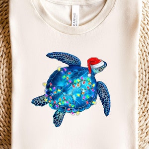 May include: A white t-shirt with a blue sea turtle wearing a red Santa hat and decorated with colorful Christmas lights.