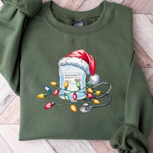 May include: Green sweatshirt featuring a graphic of a saline solution bag wearing a Santa hat, surrounded by Christmas lights and a stethoscope. The design is festive and medical-themed, perfect for healthcare workers during the holidays.