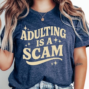 May include: Navy blue t-shirt with the text "ADULTING IS A SCAM" in a light yellow color. The text is arched and has star accents. The shirt is paired with light blue ripped jeans.