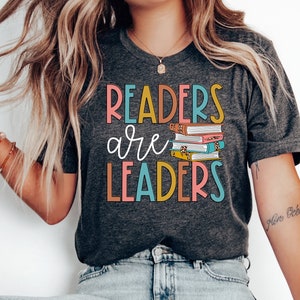 May include: A gray t-shirt with the text "Readers are Leaders" in colorful letters. A stack of books with colorful covers is pictured next to the text.