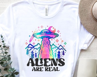 May include: White t-shirt featuring a colourful graphic of a UFO, mountains, and the text "Aliens Are Real".