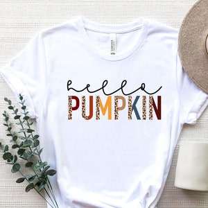 May include: White t-shirt with the text "hello pumpkin" in a colourful, leopard print design. The shirt is laid flat on a white surface with a hat and a mug.