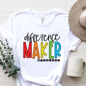 May include: White t-shirt with the text "difference maker" in rainbow colors and the hashtag "#teacher" in black.