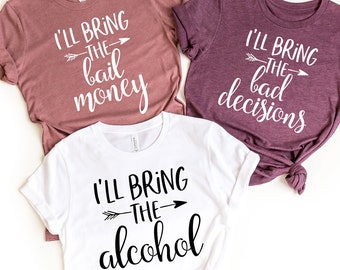 May include: Three t-shirts with different sayings. The first shirt is a dusty rose colour with the text "I'll bring the bail money". The second shirt is a dark purple colour with the text "I'll bring the bad decisions". The third shirt is white with the text "I'll bring the alcohol".