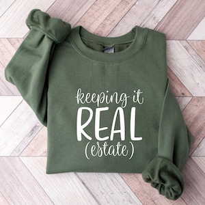 May include: A green sweatshirt with the text "keeping it REAL (estate)" in white.