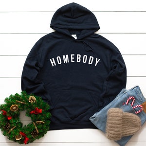 Peut inclure: Un sweat-shirt &agrave; capuche bleu marine avec le mot "HOMEBODY" imprim&eacute; en blanc sur le devant. Le sweat-shirt est pos&eacute; &agrave; plat sur une surface en bois blanc avec une couronne de verdure, un jean bleu et un bonnet marron.