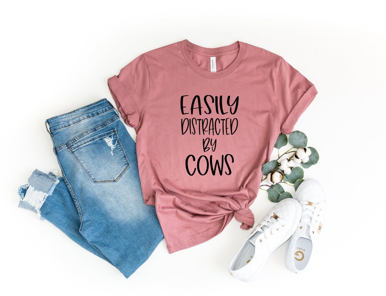 Pu&ograve; includere: Una maglietta rosa polverosa con il testo "Easily Distracted by Cows" in lettere nere. La maglietta &egrave; abbinata a jeans blu e scarpe da ginnastica bianche.