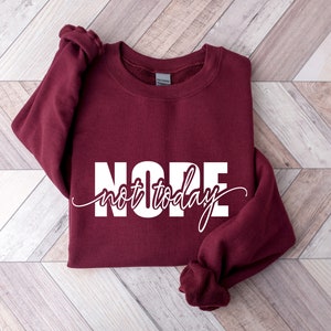 Puede incluir: Una sudadera burdeos con la palabra "NOPE" en letras blancas grandes, con "not today" en una fuente cursiva debajo. La sudadera est&aacute; doblada sobre una superficie con estampado de chevr&oacute;n marr&oacute;n claro y blanco.