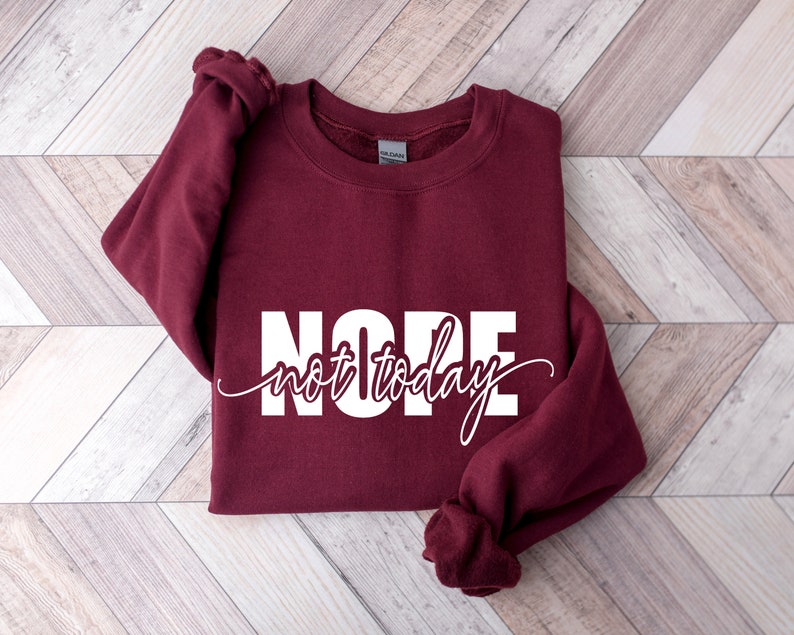 Puede incluir: Una sudadera burdeos con la palabra "NOPE" en letras blancas grandes, con "not today" en una fuente cursiva debajo. La sudadera est&aacute; doblada sobre una superficie con estampado de chevr&oacute;n marr&oacute;n claro y blanco.