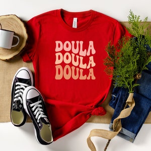May include: A red t-shirt with the word "DOULA" in a retro font, repeated three times in shades of pink and peach. The shirt is styled with black sneakers, blue jeans, and a white mug.