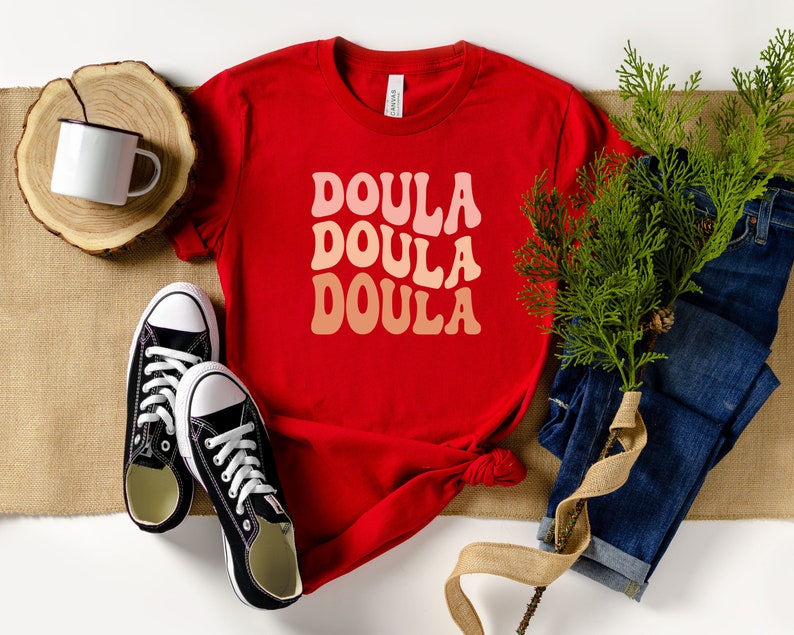 May include: A red t-shirt with the word "DOULA" in a retro font, repeated three times in shades of pink and peach. The shirt is styled with black sneakers, blue jeans, and a white mug.