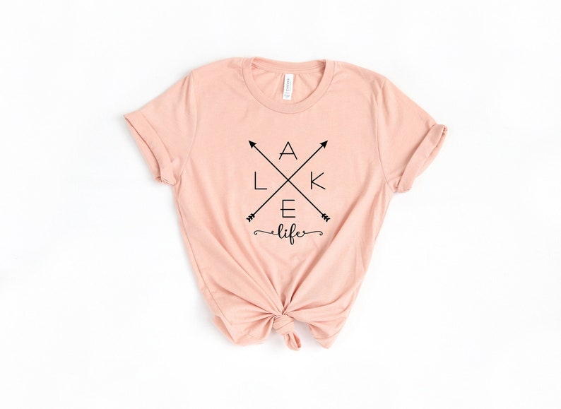 Op de afbeelding: Een lichtroze t-shirt met de woorden "LAKE life" in het zwart. Het woord "LAKE" is in een kruisvorm met pijlen gerangschikt. De shirt is aan de onderkant geknoopt.