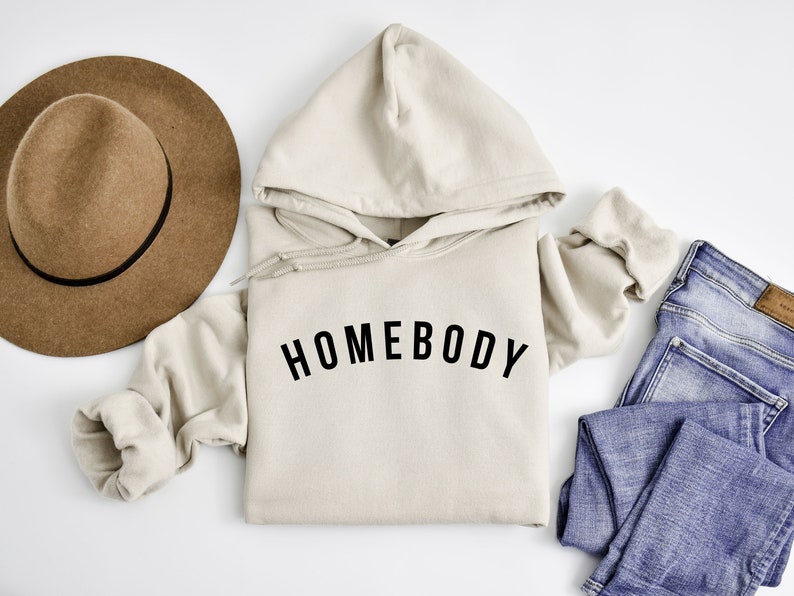Peut inclure: Un sweat &agrave; capuche beige avec le texte "HOMEBODY" imprim&eacute; en noir sur le devant. Le sweat est pos&eacute; &agrave; plat sur une surface blanche avec un chapeau marron et un jean bleu &agrave; c&ocirc;t&eacute;.