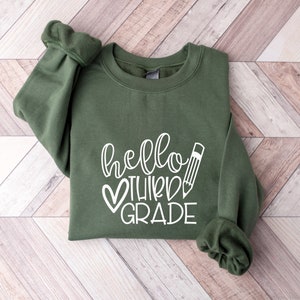 Puede incluir: Una sudadera verde con el texto "hello third grade" en letras blancas con un coraz&oacute;n y un gr&aacute;fico de l&aacute;piz.