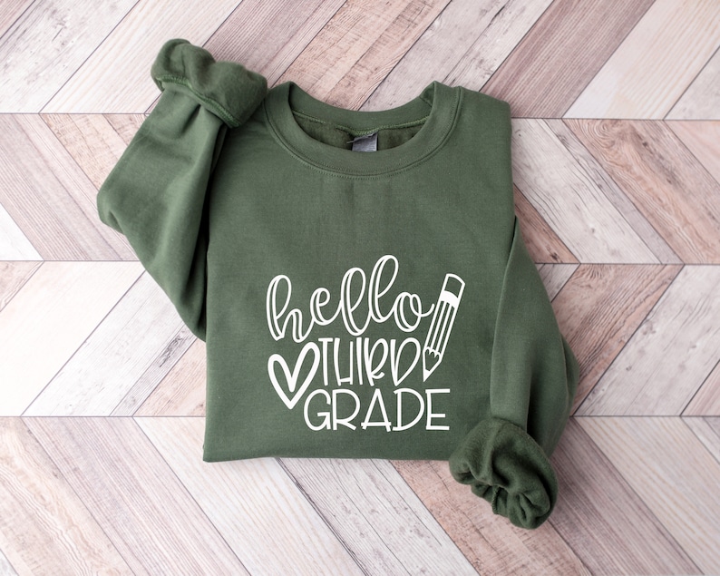 Puede incluir: Una sudadera verde con el texto "hello third grade" en letras blancas con un coraz&oacute;n y un gr&aacute;fico de l&aacute;piz.