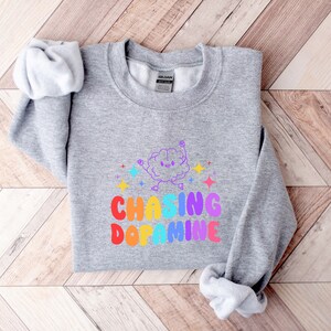 May include: A gray sweatshirt with a rainbow graphic that says "Chasing Dopamine". The graphic features a cartoon brain with a happy face and stars.
