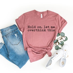 May include: A dusty rose t-shirt with the text "Hold on, let me overthink this" printed in black. The shirt is paired with blue jeans and white sneakers.