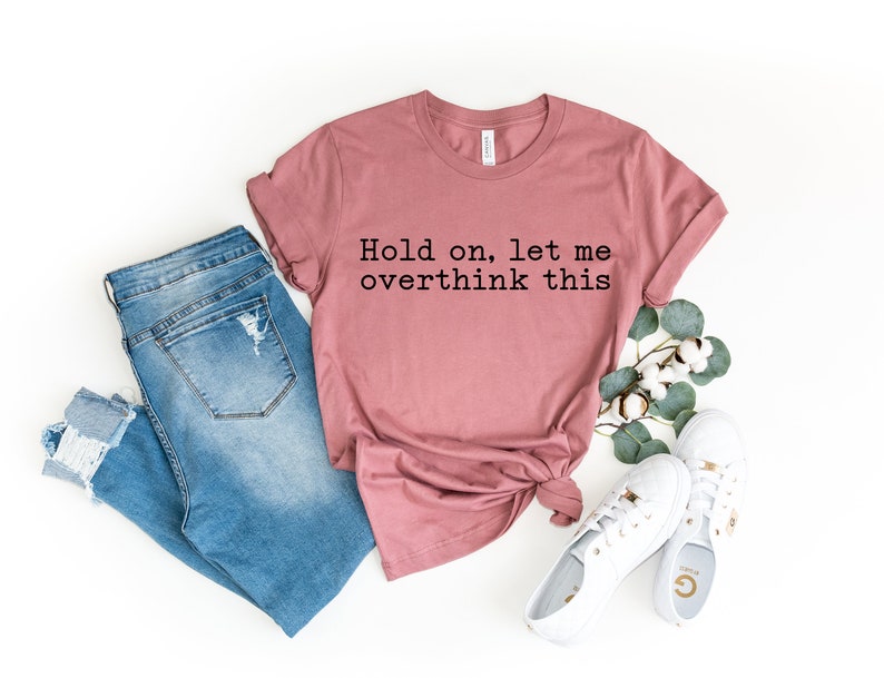 May include: A dusty rose t-shirt with the text "Hold on, let me overthink this" printed in black. The shirt is paired with blue jeans and white sneakers.