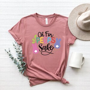 May include: A pink t-shirt with the text "Oh For Peeps Sake" in colorful letters and a graphic of a bunny with a flower.